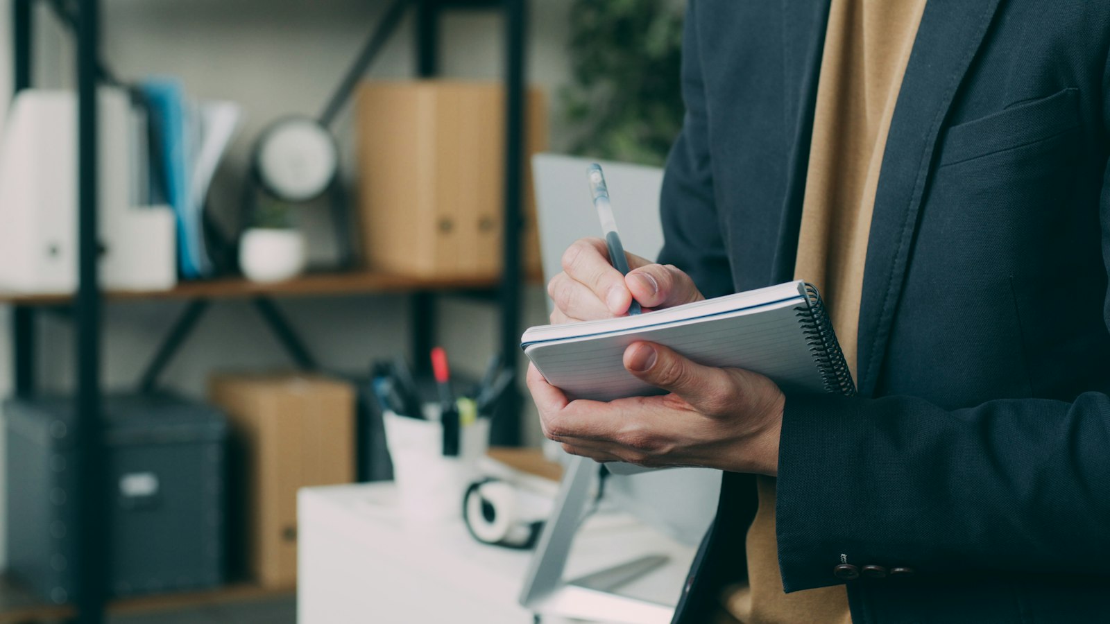 Person in a dark blazer writing notes in a notebook beside office shelves