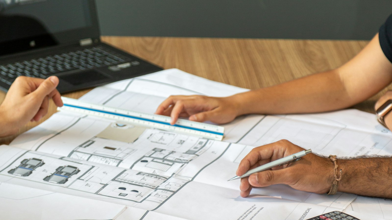 Hands pointing at architectural plans with a ruler and pen beside a laptop