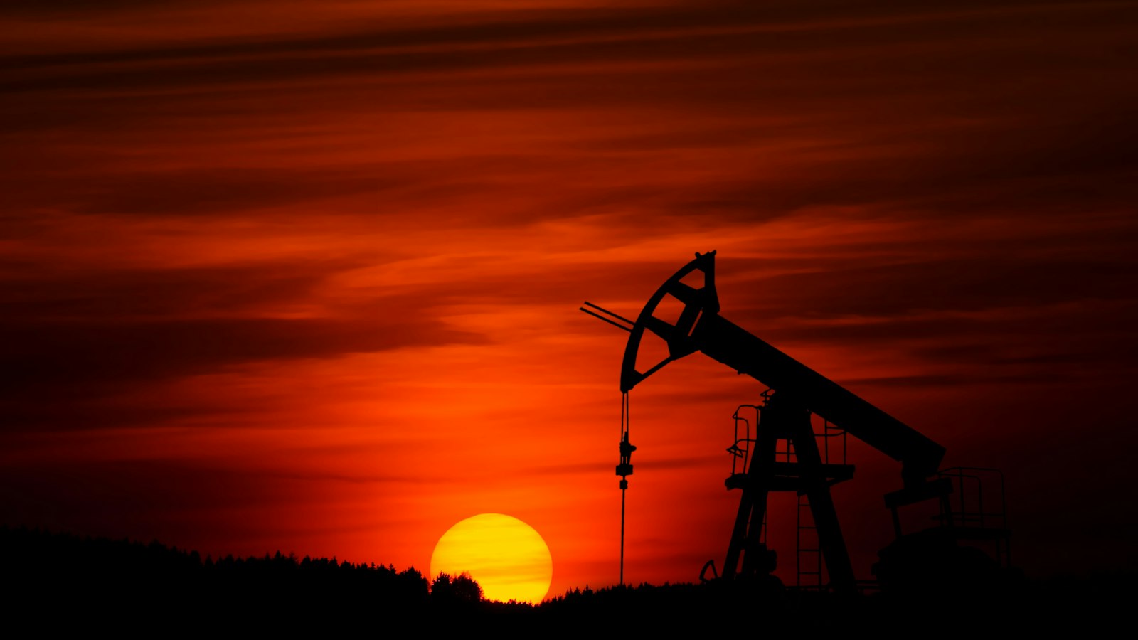 Silhouette of an oil pump jack against a deep red sunset sky