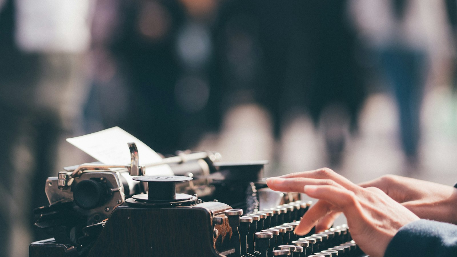 Hands typing on an old black typewriter with a blurred crowd behind