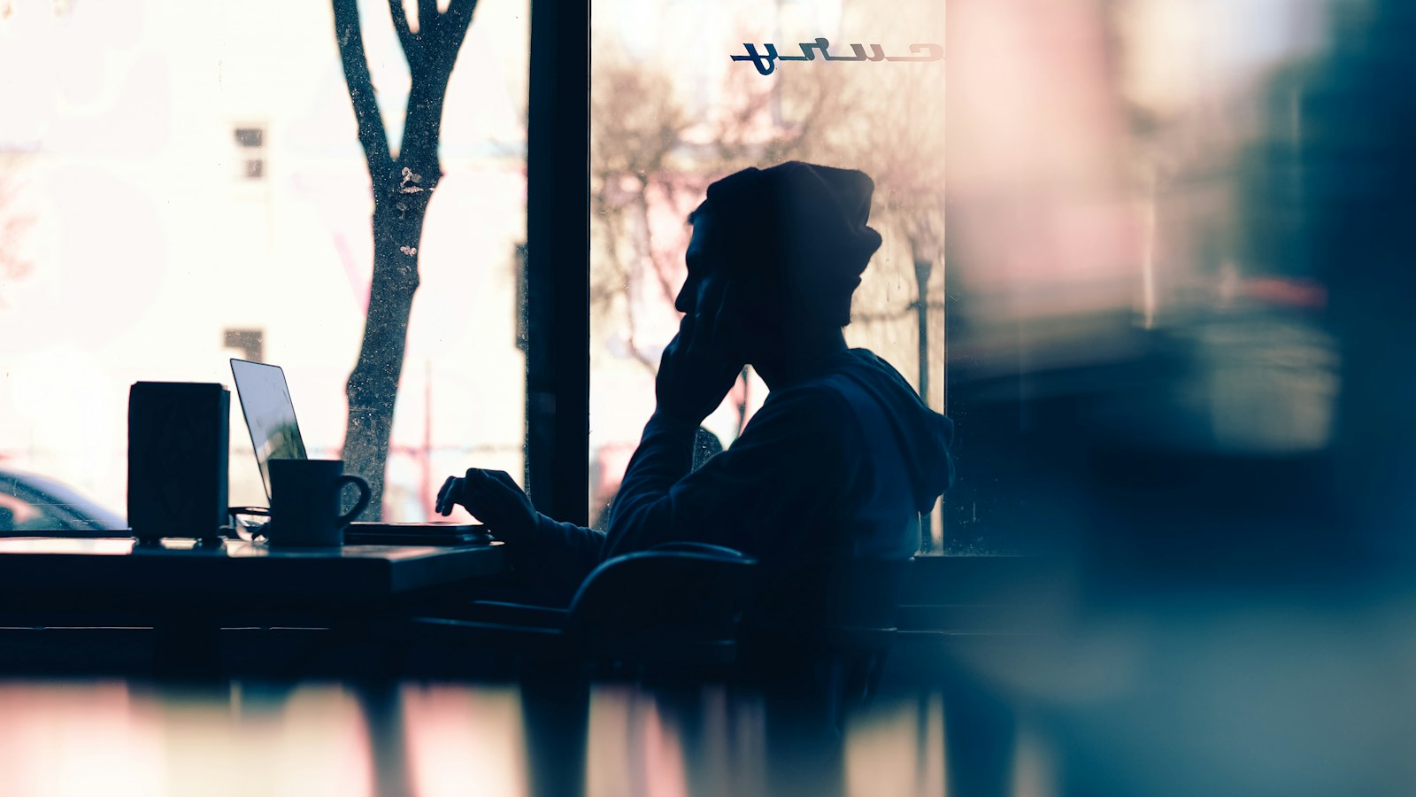 Silhouette of a person working on a laptop beside a sunlit cafe window