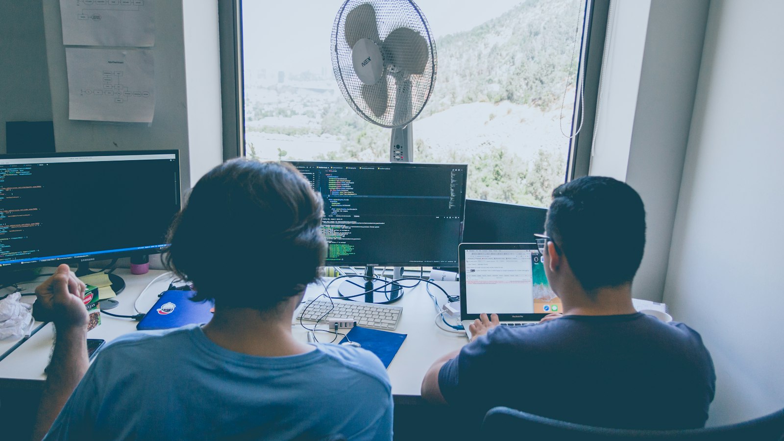 Two men seen from behind work side by side at monitors showing code
