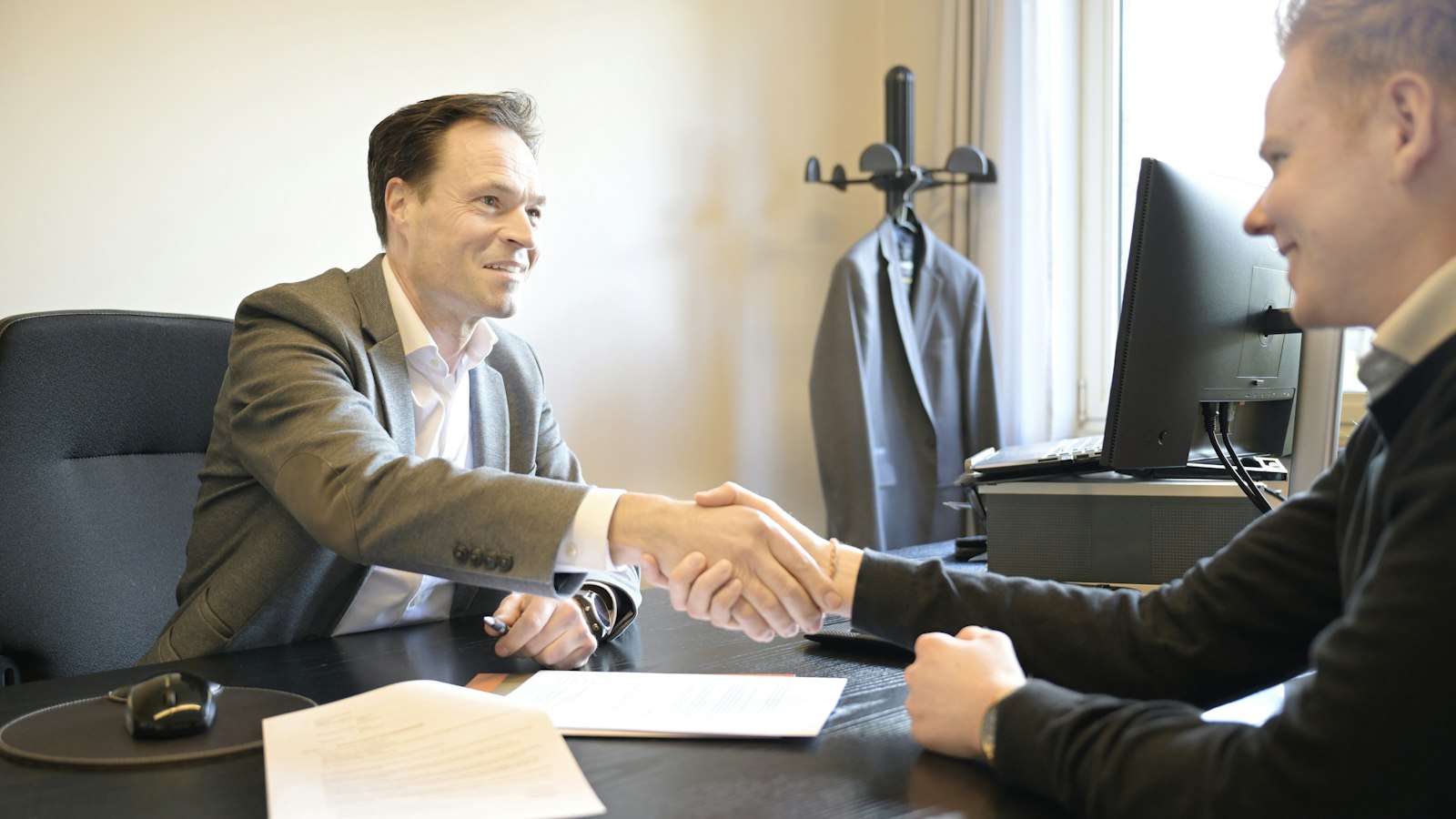 Two smiling men shake hands across an office desk over a signed document